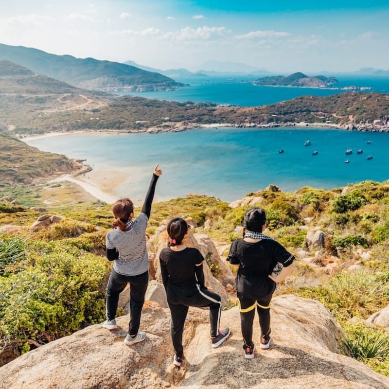 A group of three young women gazing at the landscape of the ocean from the top of a rock.