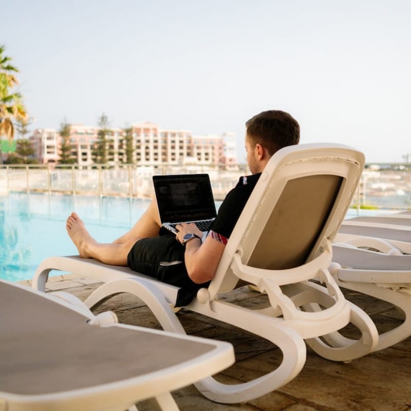 A young guy typing on his laptop, on the poolside.